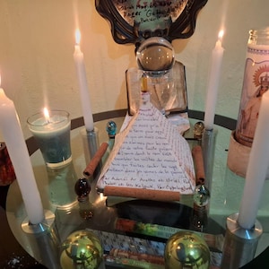 May include: A spiritual arrangement featuring lit white candles, a glass of teal liquid, and a central display with text. The arrangement includes a decorative angel figure, a crystal ball, and other decorative elements. The scene is illuminated by the candle flames.