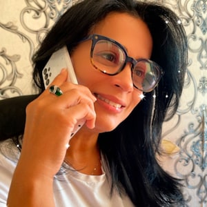 May include: A woman with dark hair is on a phone call. She is wearing a white shirt and blue-rimmed glasses with brown lenses. She is wearing a green ring on her finger.