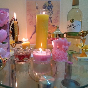 May include: A glass table top with a variety of decorative items, including a pink rose, a heart-shaped ornament, a gold bird figurine, a candle holder with a lit candle, a glass candle holder with a lit pink candle, a glass candle holder with a lit yellow candle, a glass jar with a lid, a pink candle shaped like the word "love", a gold rose with a gold base, and a bottle of white wine with the label "Carlo Rossi White".