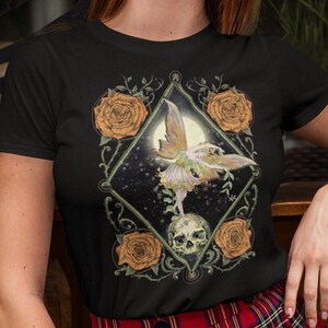 May include: Black t-shirt with a graphic print of a fairy with wings, standing on a skull, surrounded by roses and vines.