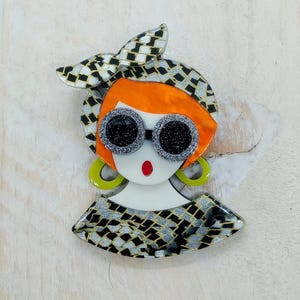 Peut inclure: Broche colorée représentant une femme stylisée avec des cheveux roux, de grandes lunettes de soleil à paillettes et un foulard et un haut à motifs. La broche a un visage blanc avec une bouche rouge et des boucles d'oreilles vertes. Le design est un mélange de noir, d'argent et d'orange.