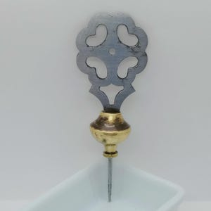 Peut inclure: Épingle décorative en métal avec un dessus ajouré argenté en forme de fleur. L'épingle a un centre bulbeux de couleur laiton et une longue épingle argentée pointue. L'épingle repose dans un récipient blanc.