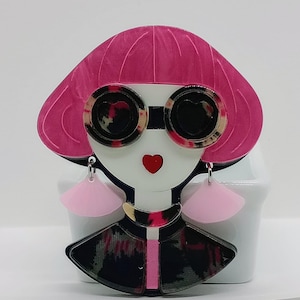 Könnte beinhalten: Eine skurrile Brosche mit einem stilisierten Gesicht mit pinkfarbenem Bob-Haarschnitt und herzförmiger Sonnenbrille. Die Brosche hat ein weißes Gesicht, einen roten herzförmigen Mund und rosa fächerförmige Ohrringe. Die Jacke ist schwarz mit einem rosa und grünen Muster.