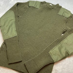 May include: An olive green ribbed knit sweater featuring solid fabric patches on the shoulders and elbows. The garment has a crew neckline, long sleeves with reinforced elbow patches, and a textured vertical knit pattern throughout the body.
