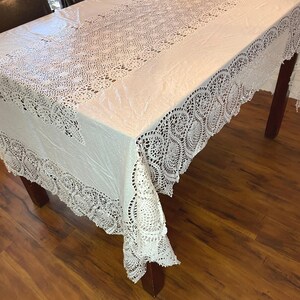 May include: A white rectangular tablecloth featuring an intricate crocheted lace border with a repeating fan-like pattern and a central panel of delicate, openwork floral crochet lace detailing that runs along the length of the table surface.