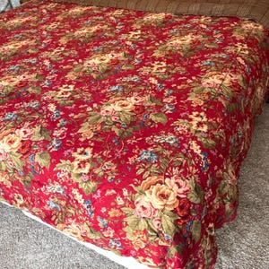May include: A quilted bedspread featuring a vibrant red background with an all-over floral pattern. The design includes clusters of roses in shades of cream, pink, and yellow, accented by green leaves and small blue flowers across the fabric surface.