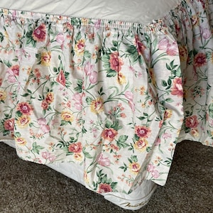 May include: A bed skirt featuring a cream-colored background with a repeating floral pattern in shades of pink, yellow, and green. The fabric has a gathered, ruffled design along the top edge, creating a soft, layered appearance around the mattress.