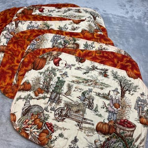May include: A set of quilted placemats featuring a harvest theme with illustrations of people in colonial-style clothing, pumpkins, corn, and wheelbarrows. The placemats have a cream-colored center with a dark orange border of autumn leaves.