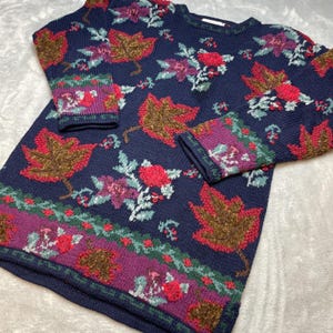 May include: Navy blue sweater with a floral and leaf pattern. The sweater features red, brown, and purple leaves and flowers. The cuffs and hem have a decorative border with similar colors and patterns. The sweater is a crew neck style.