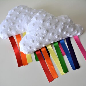 May include: Two white cloud-shaped plush toys with rainbow ribbons. The clouds have a textured surface with small, raised dots. The ribbons are in shades of red, orange, yellow, green, blue, navy, and pink.