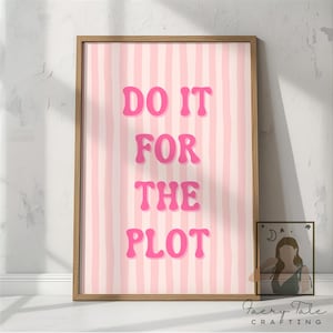 May include: A framed print featuring a pink and white striped design with the phrase "DO IT FOR THE PLOT" in pink bubble letters. The frame is a light brown colour. A smaller framed image is next to the larger print.