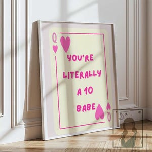 May include: A framed print with a playing card design. The text "YOU'RE LITERALLY A 10 BABE" is in pink. The card has pink heart suit symbols and a light yellow background. The frame is white.