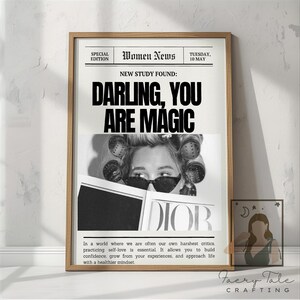 May include: A wooden framed print with a newspaper design. The headline reads "DARLING, YOU ARE MAGIC" above a black and white photo. The print includes the text "DIOR" and a quote about self-love. A smaller framed print is to the right.