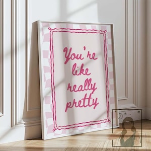 May include: A framed art print featuring the phrase "You're like really pretty" in pink script. The print has a pink and white checkered border and is propped against a white wall. The artwork rests on a wooden floor.