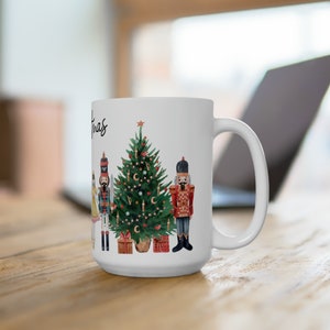 May include: A white ceramic mug featuring a watercolor illustration of a green Christmas tree, two nutcracker figures, a small doll, and wrapped gifts. The text "Christmas" is printed in black script on the side of the mug near the tree.