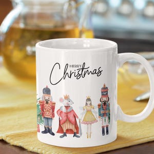 May include: A white ceramic mug featuring the text Merry Christmas in black script. The design includes a watercolor illustration of a nutcracker, a mouse king in a red cape, a fairy in a yellow dress, and another nutcracker standing in a row.