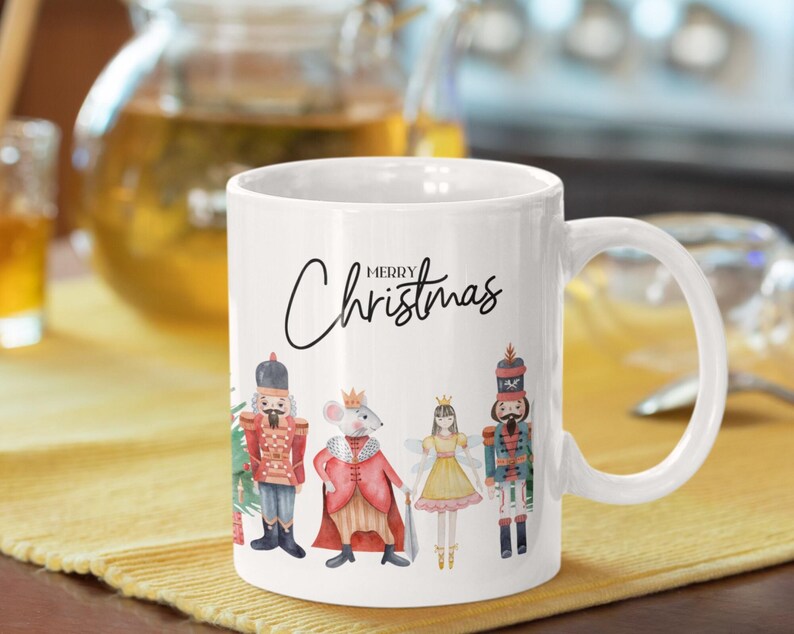 May include: A white ceramic mug featuring the text Merry Christmas in black script. The design includes a watercolor illustration of a nutcracker, a mouse king in a red cape, a fairy in a yellow dress, and another nutcracker standing in a row.