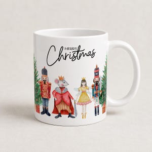May include: A white ceramic mug featuring a watercolor illustration of a nutcracker, a mouse king, a fairy, and another nutcracker standing between two Christmas trees. The text "MERRY Christmas" is printed in black script above the characters.
