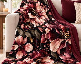 May include: A plush throw blanket featuring a dark black background adorned with large, vibrant floral patterns in shades of deep burgundy, soft pink, and cream. The fabric has a velvety texture and is draped over a light-colored sofa cushion.