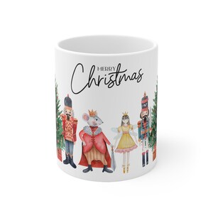 Könnte beinhalten: Weiße Keramiktasse mit einer Aquarell-Illustration eines Nussknackers, eines Mäusekönigs und einer Ballerina vor einem Weihnachtsbaum. Der Text "Merry Christmas" ist in schwarzer Kursivschrift auf der Tasse geschrieben.