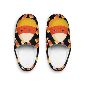 Puede incluir: Un par de pantuflas de interior negras y naranjas con un patrón de maíz dulce. El maíz dulce tiene una cara linda y está rodeado de maíz dulce más pequeño.