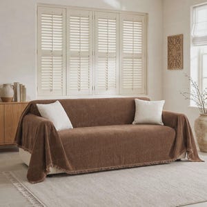 May include: A brown textured sofa cover with fringe detailing along the bottom edge, draped over a sofa and accented with two white square throw pillows. The text EmbraceComfort is visible at the bottom center of the image.