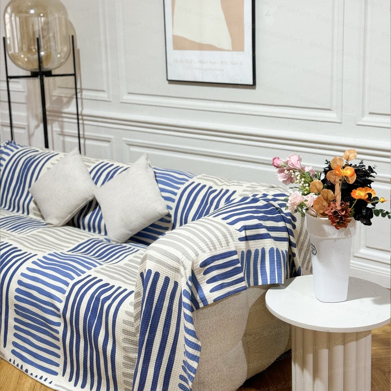 May include: A white couch with blue and gray striped throw blanket. The couch has two white pillows and is decorated with a vase of flowers.