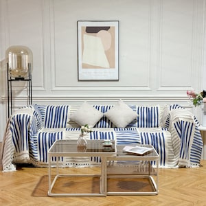 May include: A living room with a white couch with blue and beige striped throw blankets. The couch has two white throw pillows. There is a glass coffee table with a white metal frame in front of the couch. A glass floor lamp with a black metal stand is in the corner of the room. There is a framed abstract art print on the wall behind the couch.