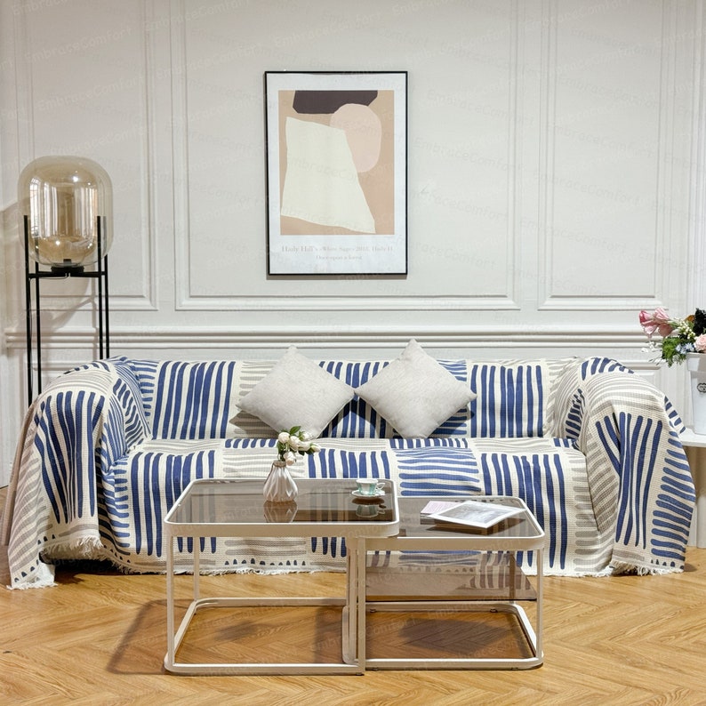 May include: A living room with a white couch with blue and beige striped throw blankets. The couch has two white throw pillows. There is a glass coffee table with a white metal frame in front of the couch. A glass floor lamp with a black metal stand is in the corner of the room. There is a framed abstract art print on the wall behind the couch.