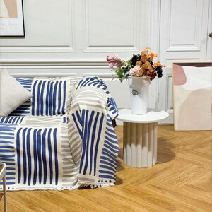 May include: A white couch with a blue and white striped throw blanket. A white side table with a white vase filled with pink, orange, and red flowers sits next to the couch.