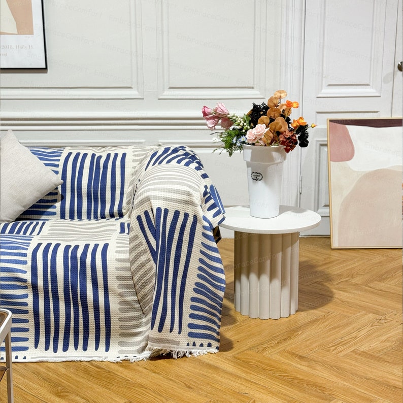 May include: A white couch with a blue and white striped throw blanket. A white side table with a white vase filled with pink, orange, and red flowers sits next to the couch.
