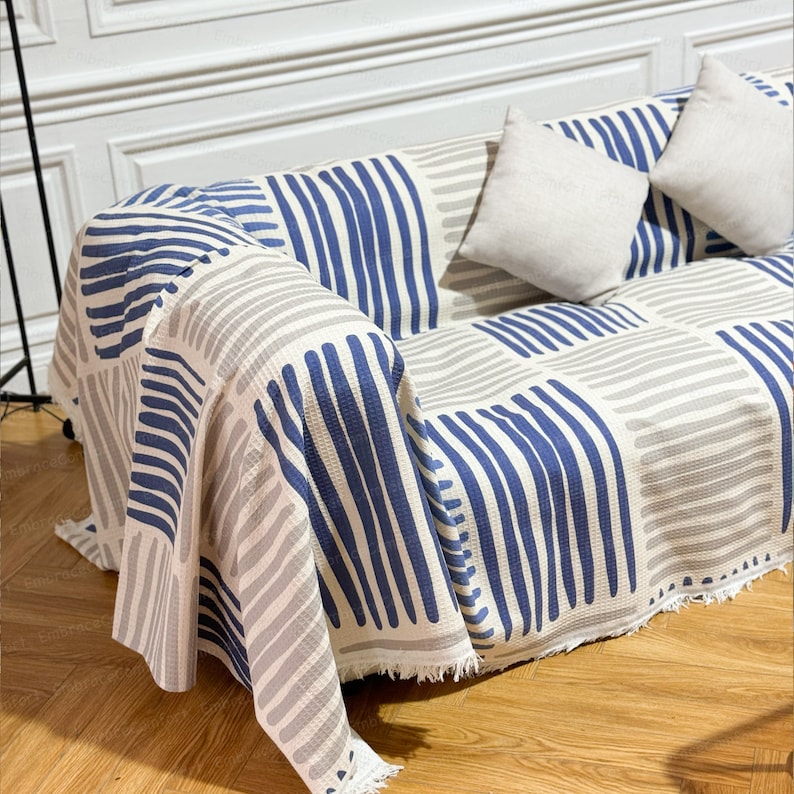 May include: A white and blue striped throw blanket with a fringe border draped over a couch. The blanket has a geometric pattern of horizontal stripes in shades of blue and gray.