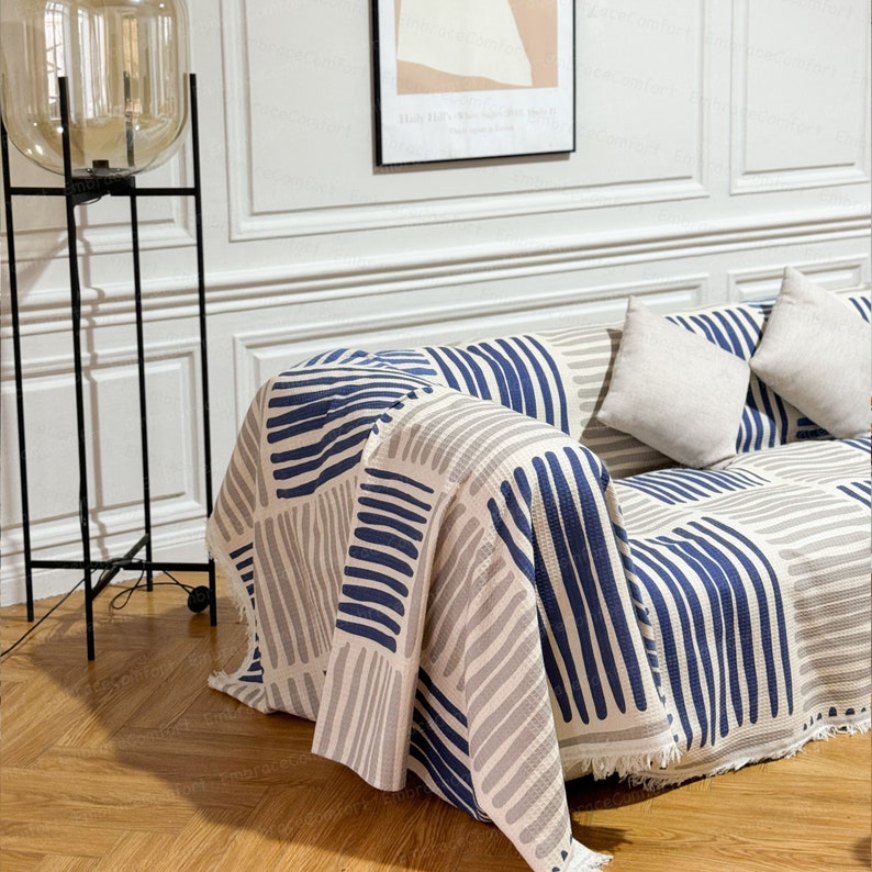 May include: A white and blue striped throw blanket with fringe edges draped over a couch. The blanket has a geometric pattern of thick and thin stripes.