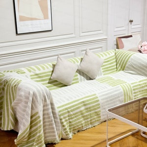May include: A green and white striped throw blanket with fringed edges draped over a sofa with two white cushions. The sofa is in a living room with a wooden floor and a glass coffee table.