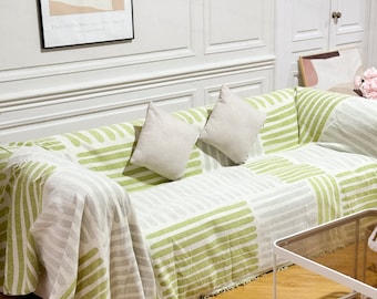 May include: A green and white striped throw blanket with fringe edges draped over a couch with two white pillows. The couch is in a living room with a wooden floor and a glass coffee table.