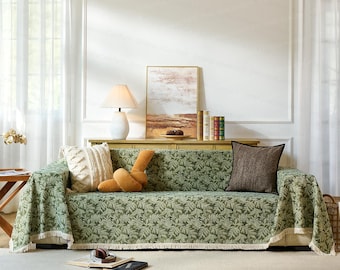May include: A green sofa with a leaf pattern is covered with a matching throw. Decorative pillows in cream, brown, and tan are arranged on the sofa. A lamp, artwork, and books are on a wooden console behind the sofa. The text "EmbraceComfort" is at the bottom.