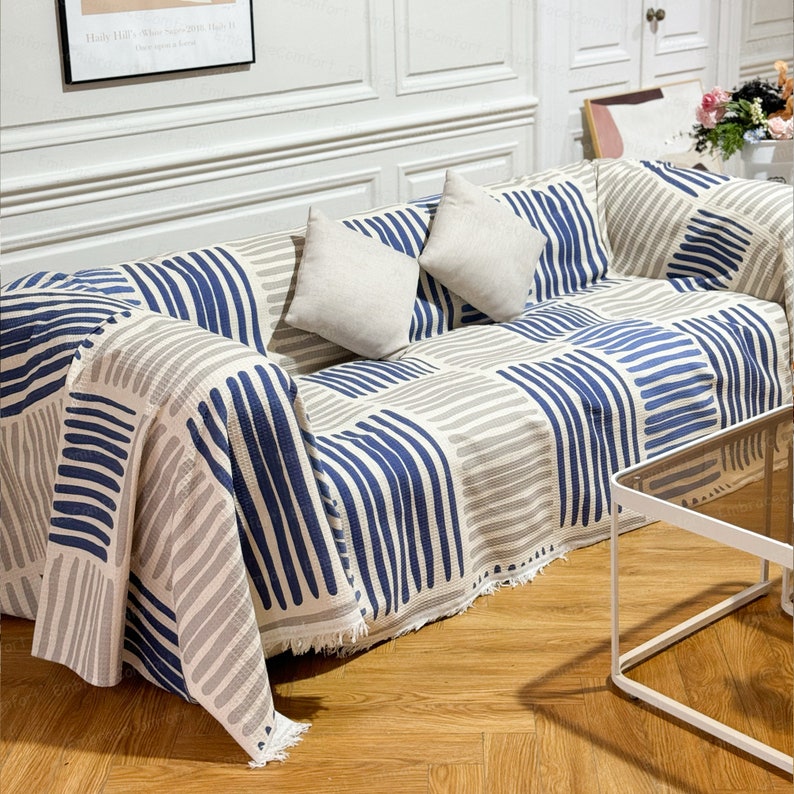 May include: A white and blue striped throw blanket with a fringe edge. The blanket is draped over a couch with two white throw pillows.