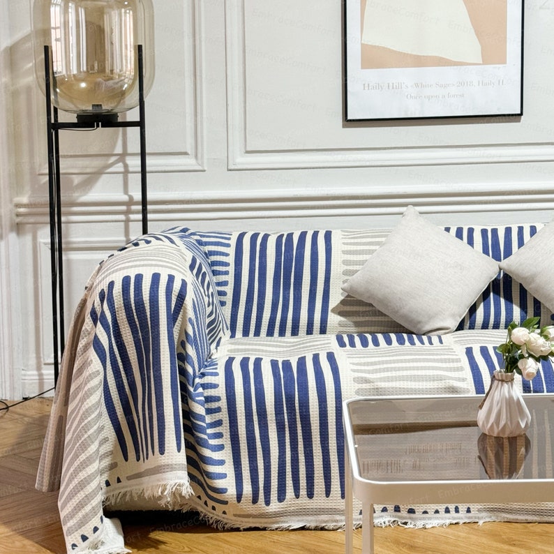 May include: A white and blue striped throw blanket with a fringe edge draped over a couch. The couch has two white pillows and a small white table with a glass top in front of it.