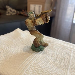 May include: A small, detailed figurine of Chewbacca from Star Wars, holding a blaster. The figure is primarily brown and tan, with a green base. The blaster is a light tan color. The figurine is posed in a dynamic action stance.