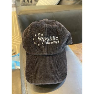 May include: A dark brown corduroy baseball cap with the text "Republic Airways" embroidered in white. The cap has a curved brim and a structured crown. The text is accompanied by small star designs.