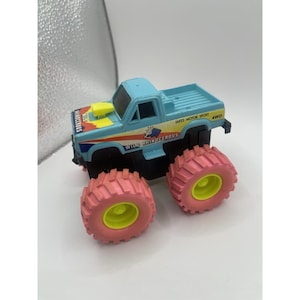 May include: A light blue toy monster truck with pink tires and yellow accents. The truck has the words "Wild Rhinoceros" and "Jakes Motor Sport 4WD" printed on it. A fun toy for kids.