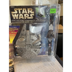 May include: A Star Wars Collector Timepiece set, including a Millennium Falcon watch case and a Darth Vader watch. The packaging includes the Star Wars logo and text that reads "Collector Timepiece". The watch has a black band.