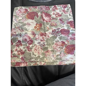 May include: A square fabric napkin featuring a dense floral pattern with shades of deep red, burgundy, soft pink, and muted sage green on an off-white background. The design displays various blooming flowers and green leaves across the surface.