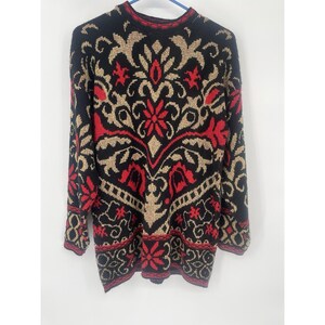 May include: A long-sleeved sweater with a black background and an intricate pattern in red and tan. The pattern includes floral and geometric shapes, with red accents on the collar and cuffs. The sweater is hanging on a blue hanger.
