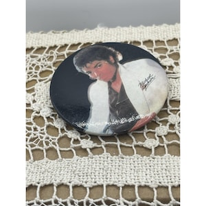 May include: A round button featuring a portrait of Michael Jackson in a white jacket against a black background. The button has the name "Michael Jackson" printed on it.