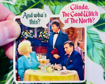 Peut inclure: Un autocollant rectangulaire représentant une scène de restaurant avec trois personnages. Des bulles de dialogue indiquent "And who's this?" et "Glinda, The Good Witch of The North?". L'autocollant a une bordure blanche et est tenu par une main.