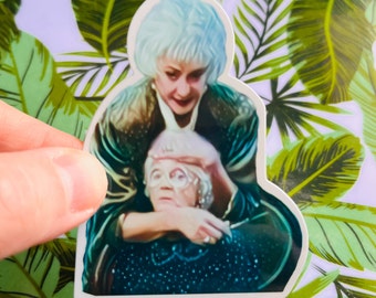 May include: A sticker featuring a close-up of two women, one with white hair and a blue jacket, the other with gray hair and a blue dress. The woman with white hair is holding the other woman's face in her hands.