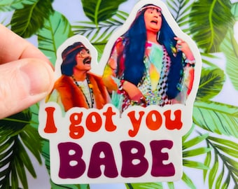 Könnte beinhalten: Ein Aufkleber mit zwei Personen in Kleidung aus den 1970er Jahren mit dem Text "I got you babe" in Orange und Lila.