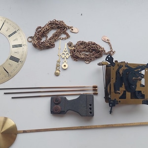 May include: Clock parts including a gold-colored clock face with black Roman numerals, a pendulum, chains, and a mechanical movement. The components are arranged on a white surface, suggesting a restoration project or a collection of vintage timekeeping elements.