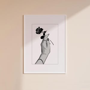 Op de afbeelding: Zwart-wit kunstafdruk van een hand die een zwarte roos vasthoudt. De hand is versierd met ringen en een armband. Het kunstwerk is ingelijst in een witte rand, wat een minimalistische esthetiek creëert.